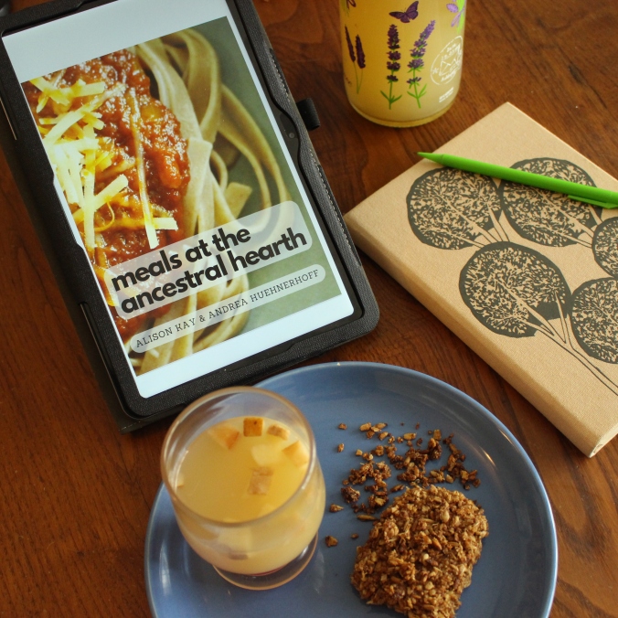 picture of an ereader with a cookbook open in it, with the words Meals at the Ancestral Hearth. A plate with fermented ancestral drink and a grassmere gingerbread slice is sitting next to it. There is a swing-top bottle with more water kefir, and a notebook and pen for taking notes. 